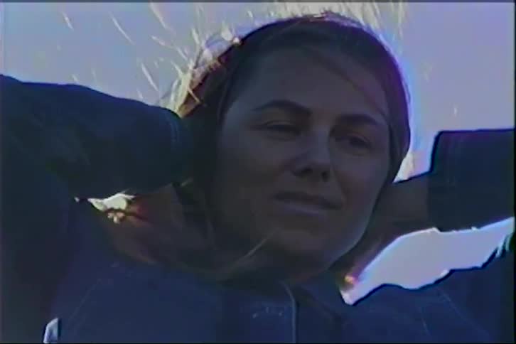 A woman with long hair stands under a clear blue sky smiling down at the camera and running her hands through her windblown hair while wearing a denim jacket in a lowangle shot