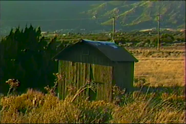 A small hut sits on a wide grass field in the countryside at midday with a distant mountain range rising behind it