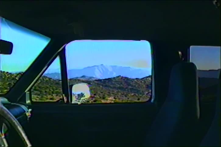 From inside a parked vehicle a sunlit grassylandscape and distant mountains are framed through the side window