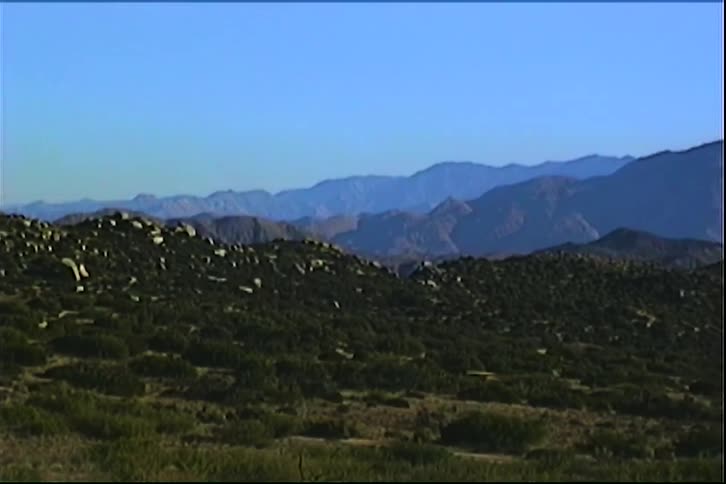 A wide static shot captures a vast desert landscape featuring green scrubcovered hills in the foreground and a massive bluetinted mountain range under a clear sky during the day