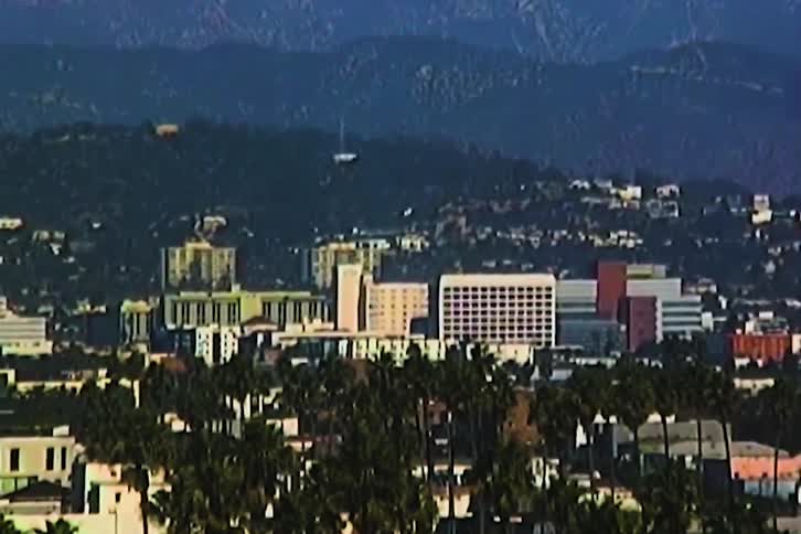 A sprawling urban skyline nestled against a backdrop of hazy mountains with residential rooftops and palm trees filling the foreground under a bright clear sky during the day