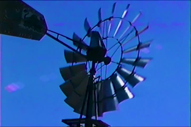 A vintage metal windmill silhouetted against a clear saturated blue sky its circular blades and tail fin catching the light as they arc outward