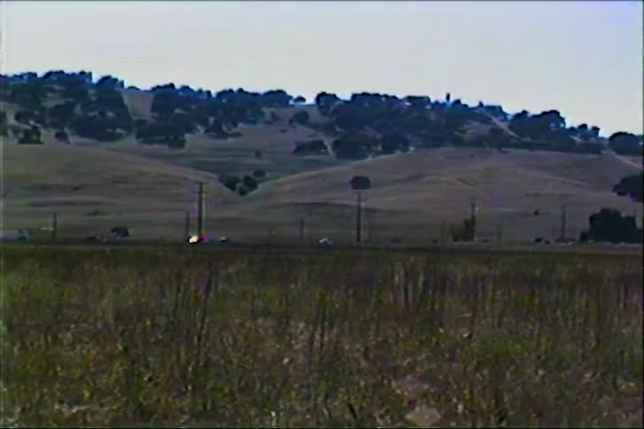 A line of cars travels along a highway that stretches across a vast goldenhued valleyin the background rolling hills dotted with sparse trees rise under a clear bright sky captured with a vintage vhs aesthetic