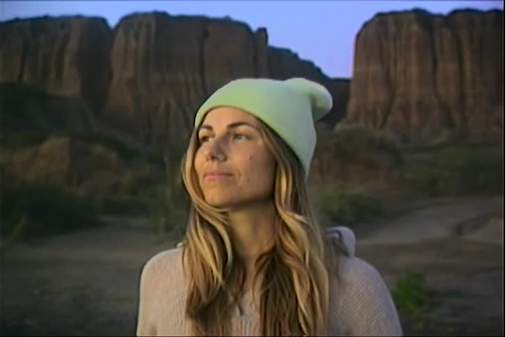 A young woman stands in the wilderness at midday smiling as she hikes with a mountain rising behind her her long hair flowing beneath a warm beanie hat