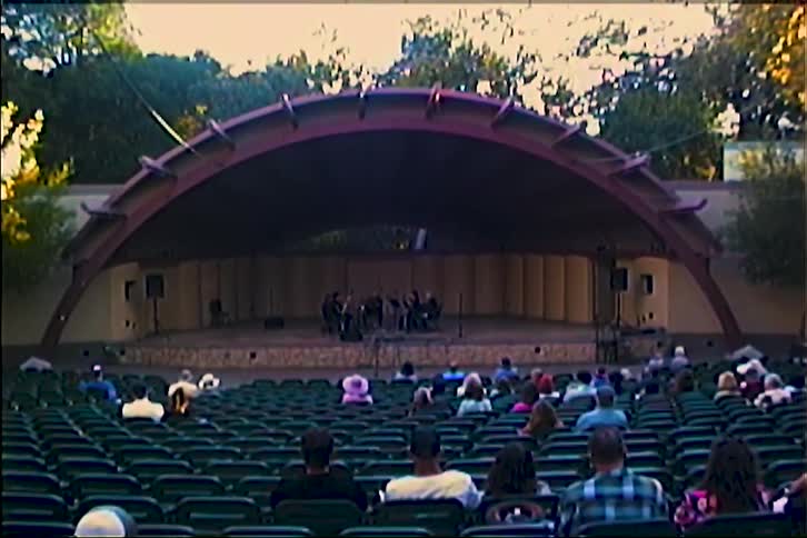 An audience sits in an outdoor amphitheater watching a musical ensemble perform on a stage under a large curved shell the scene shows rows of green seats filled with scattered spectators as a small group of musicians plays in the center of the stage
