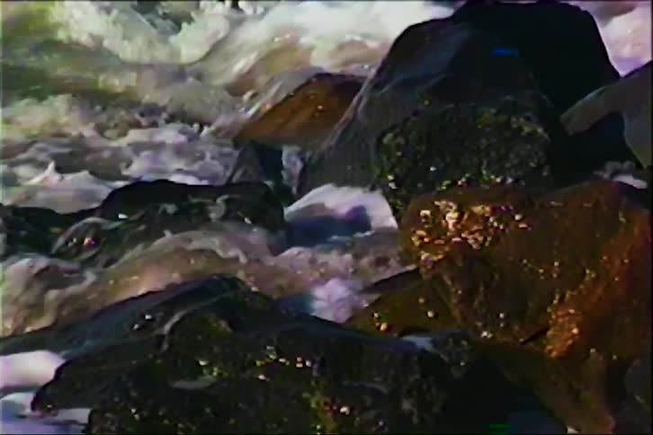 Foamy fastflowing water rushes past dark wet rocks in a grainy vintagelooking frame with swirling white currents contrasting against the shadowed stone surfaces