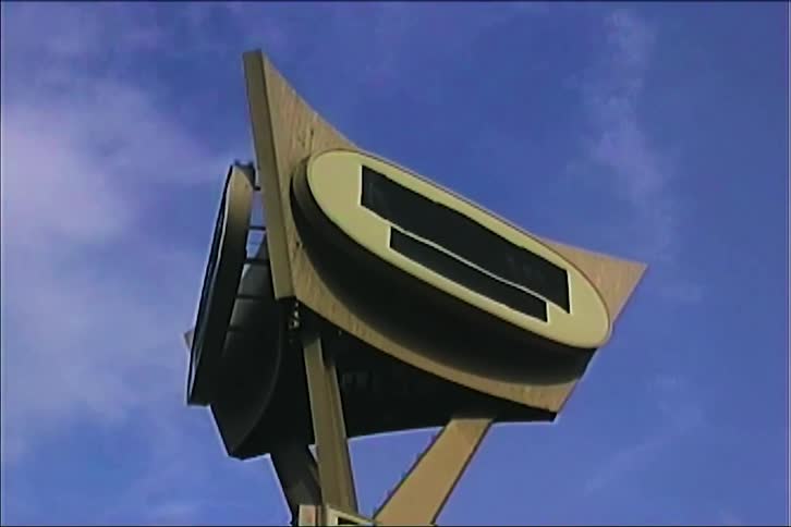 A vintage architectural structure with geometric gold frame and blank oval center sits atop a metal support against a clear blue sky with light clouds