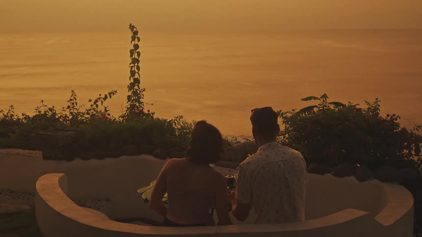 Two people seated from behind on a terrace with a set table overlooking the sea under golden dusk light