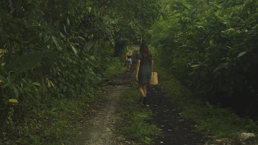 Three women walk down a narrow shaded dirt path through a lush green forest