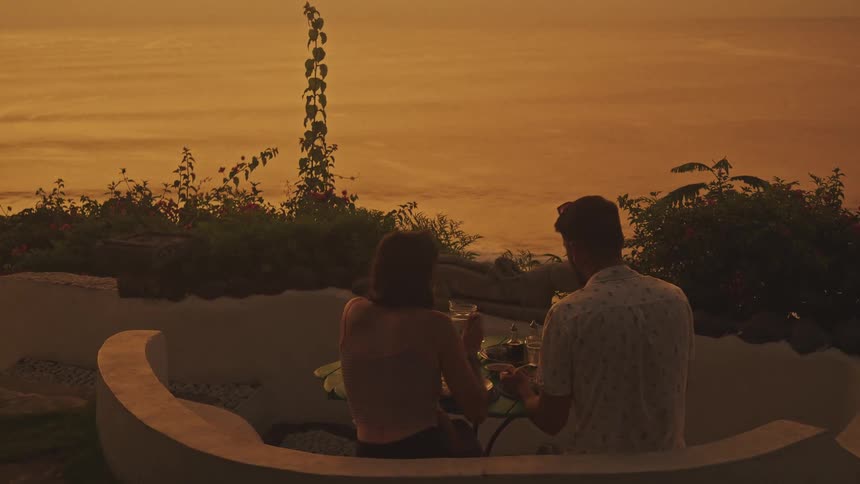 A couple enjoys a romantic meal together on a patio silhouetted against the golden glow of the ocean at sunset