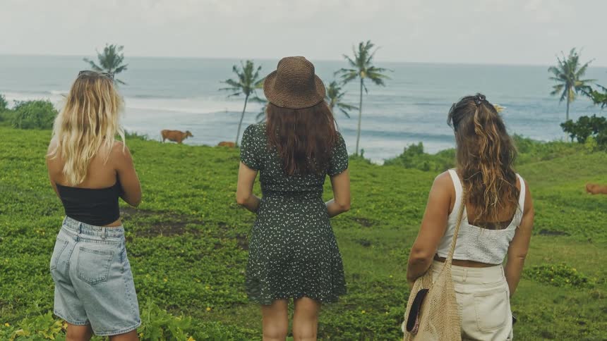 Three women stand in a field overlooking the ocean under an overcast sky
