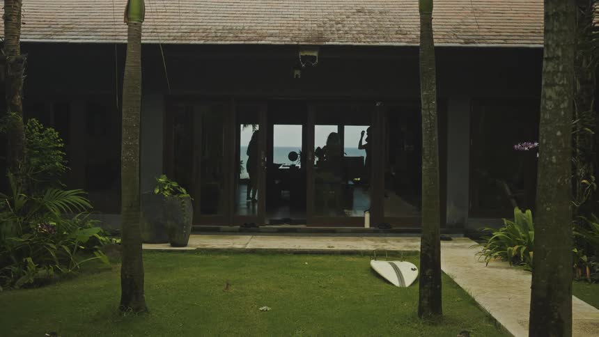 A house with glass doors overlooks a beach where silhouettes of people are seen inside outside a grassy yard contains palm trees and a white surfboard lying on the ground