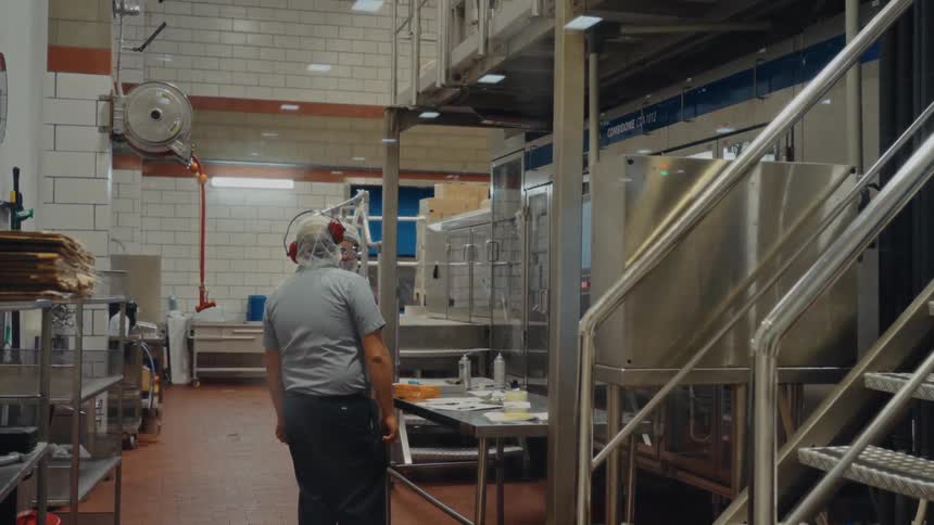 In a sterile industrial kitchen a worker wearing protective headgear and a hairnet stands near large stainless steel machinerythe facility features tiled walls and organized workstations emphasizing a clean and professional food production environme