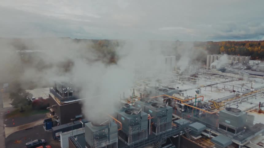 An aerial shot pulls back from a large factory revealing its complex rooftop where thick white steam billows from numerous industrial vents