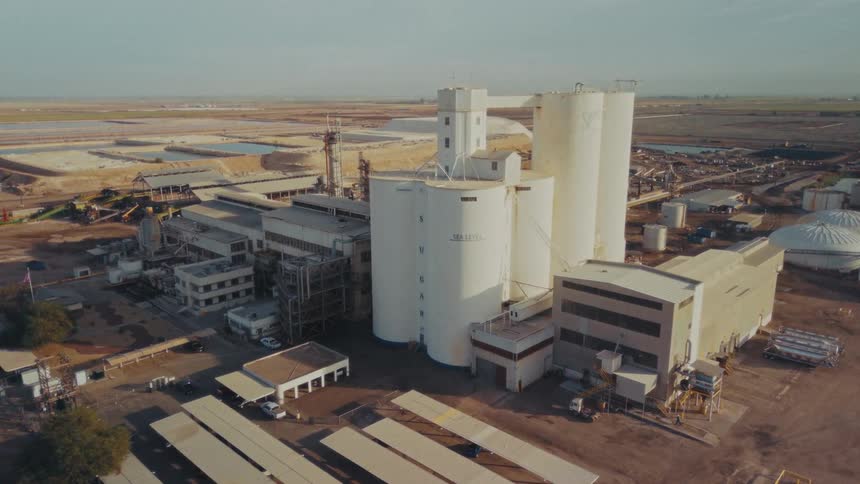 An aerial drone shot pans over a large white industrial processing facility and tall silos situated in a flat desert environment featuring evaporation ponds and vast dirt fields under a clear sky
