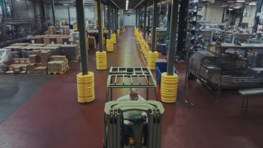 A forklift driver wearing a white shirt and safety glasses navigates through a wide aisle in a busy industrial warehousethe facility is organized with rows of structural columns protected by yellow safety padding and various pallets of boxed goods sta