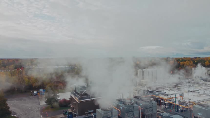 Industrial facility with towers and pipelines releasing steam in an area surrounded by vegetation