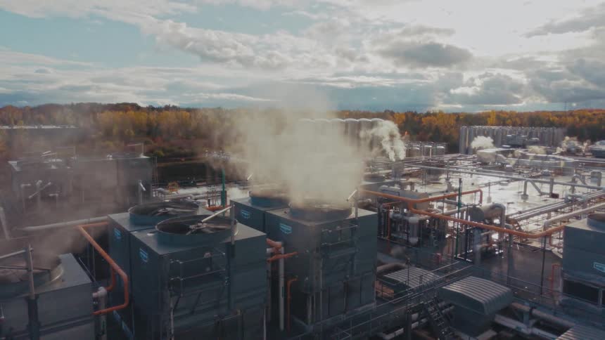 An aerial camera pushes in slowly over a sprawling factory rooftop where thick white steam billows from large industrial cooling towers and vents
