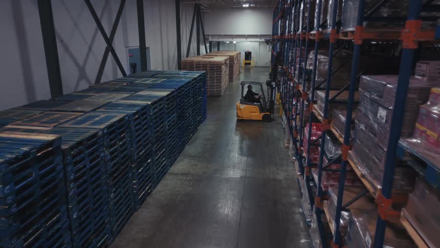 Forklift operating in a warehouse aisle between tall racks with pallets and stored goods