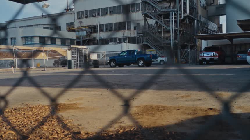View through a chain link fence of trucks in a parking lot and a factory building with outside stairs