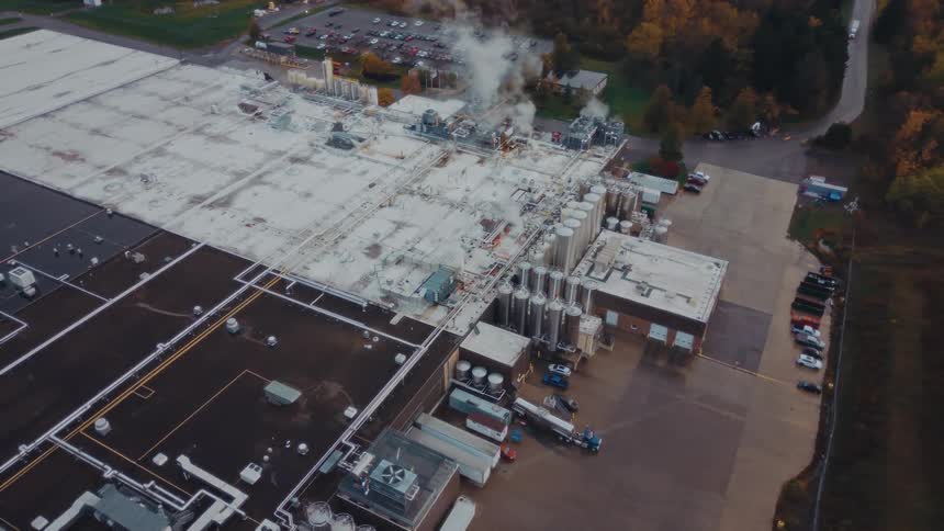 Aerial view of a factory with steaming vents amidst autumn trees and parking lots