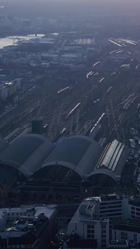 An aerial view shows a massive railway station with vaulted roofs and numerous tracks extending into a hazy urban horizon at dusk as small trains move through the industrial cityscape near a body of water and surrounding apartment blocks