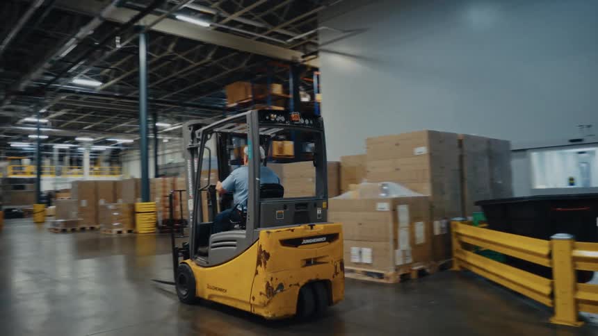 A male warehouse worker wearing a blue shirt and gray cap drives a yellow forklift through a large distribution center filled with stacked cardboard boxes on pallets passing yellow safety railings and other parked machinery