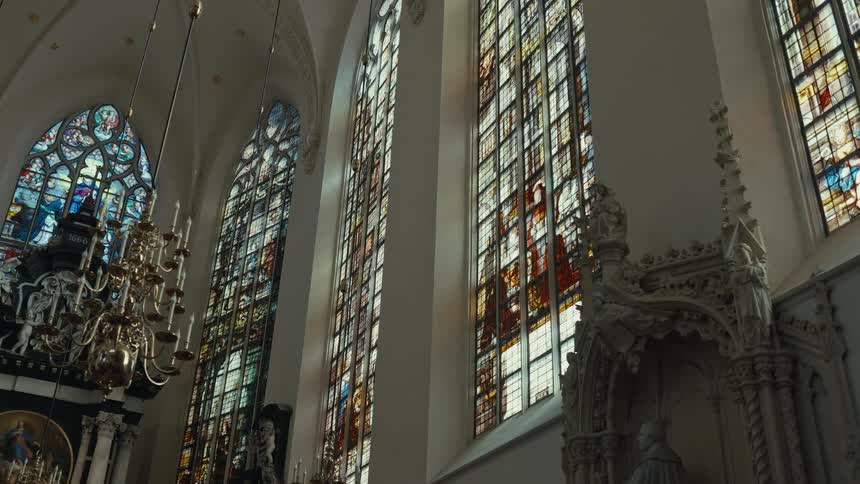 A camera pans slowly across the interior of a church showcasing tall gothic windows filled with intricate stained glass art while a brass chandelier and ornate stone sculptures sit in the foreground and lower corners of the whitewalled space