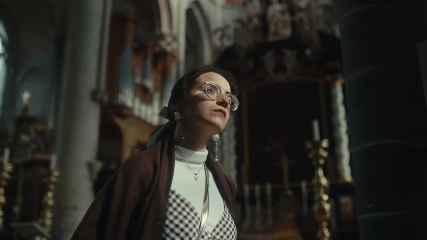 A teenage girl inside a castle wearing glasses and a brown jacket curiously examining the antiques in the building