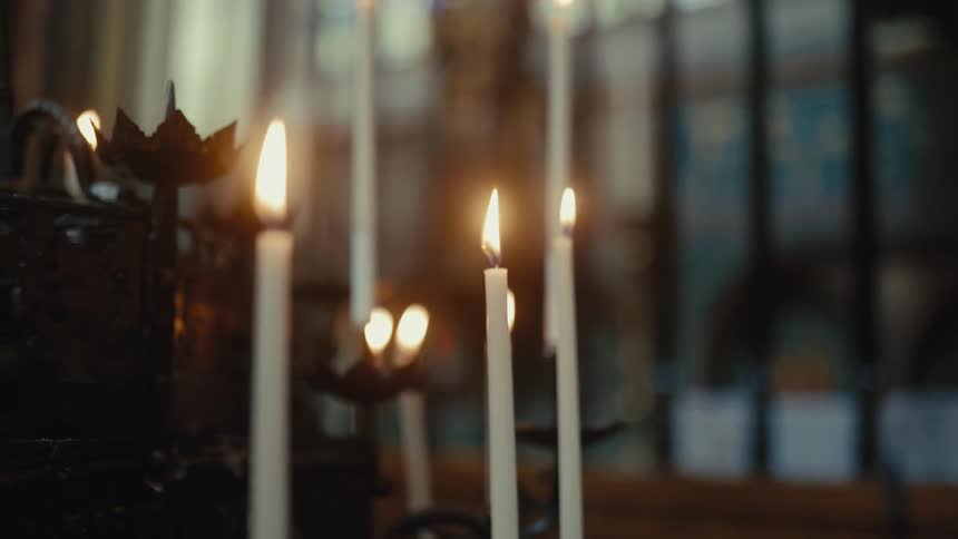 A group of tall white taper candles burning in a dimly lit somber environment that appears to be the interior of a church or cathedral as the camera slowly zooms in the warm glow of the flickering flames creates a sharp contrast against the blurred