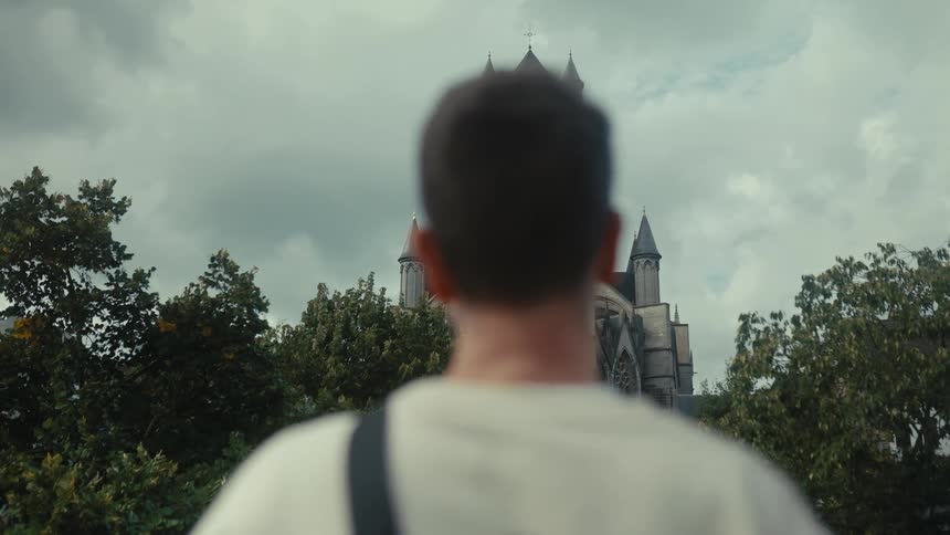 Person seen from behind looks toward a gothicstyle church with tall towers above the treetops under a cloudy sky