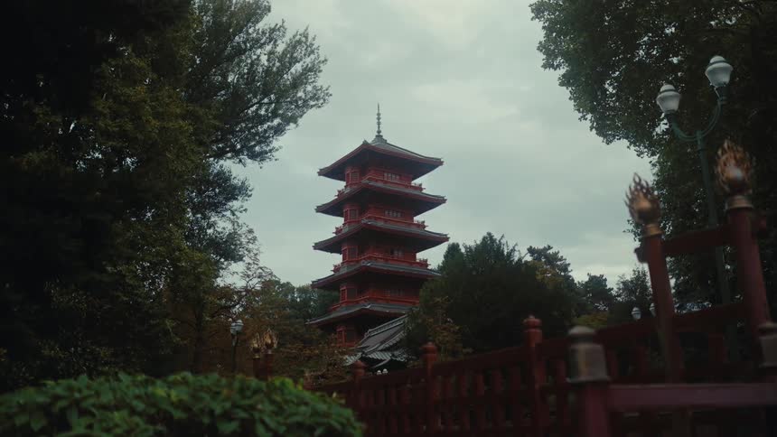 A red multistory pagoda stands tall among lush green trees under an overcast sky a decorative wooden fence and a vintagestyle lamp post frame the foreground of the serene landscape