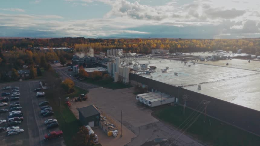 Aerial view of a factory complex with steam rising surrounded by autumn trees and parking lots