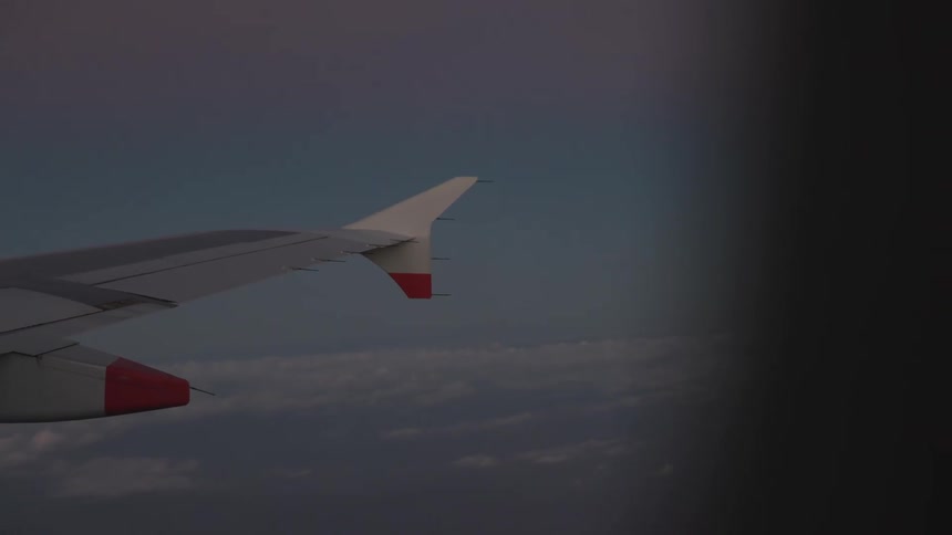 An airplane wing stretches across a muted blue sky above a soft blanket of clouds with red engine accents and a dark outoffocus edge