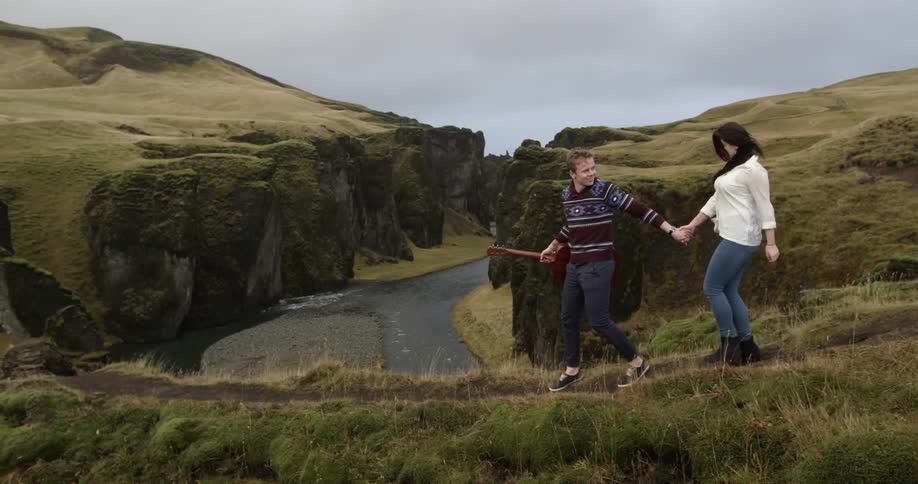 Woman and man with guitar holding hands are walking down mountain path