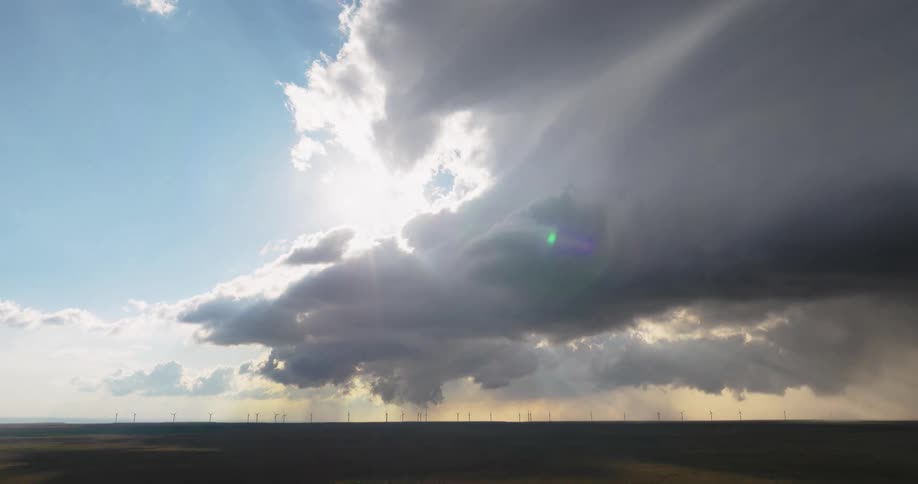 In a dramatic timelapse a massive storm system sweeps over a vast wind farm as powerful sun rays pierce through the dark swirling clouds
