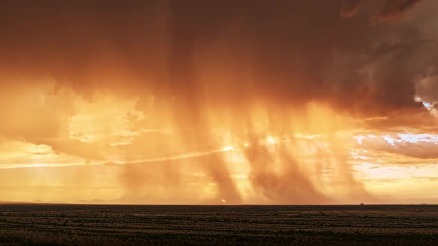 A vast flat landscape stretches beneath a dramatic sky as towering storm clouds release golden curtains of rain backlit by a low setting sun that glows intensely at the horizon