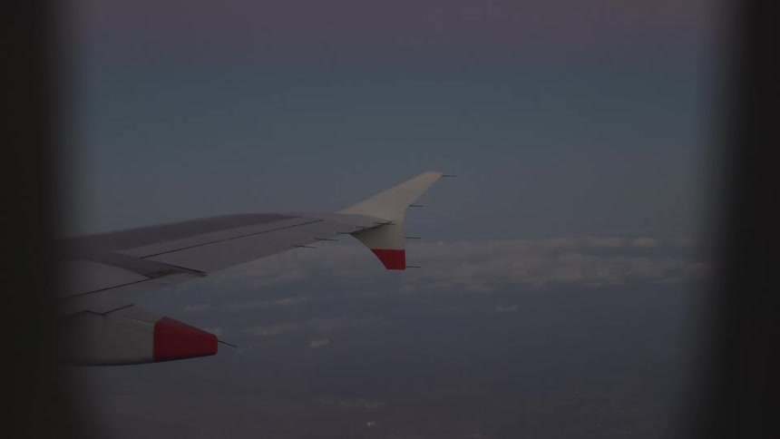 An airplane wing with distinctive redtipped engines and winglets is visible through a passenger window as it flies over a vast landscape dense layers of white clouds stretch across the horizon beneath a darkening purple and blue twilight sky