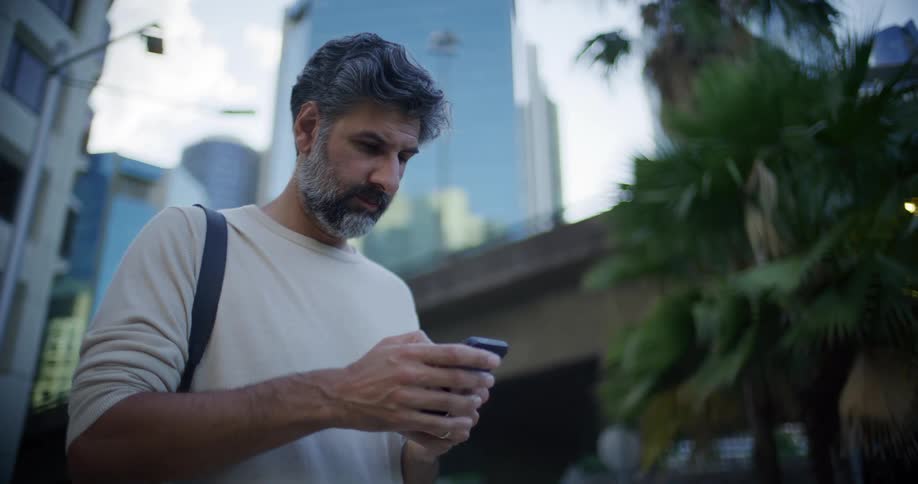 A middleaged man with graying hair and a beard stands on an urban street while focused on his smartphone typing with his thumbs against a backdrop of city buildings a concrete overpass and green foliage under a clear sky
