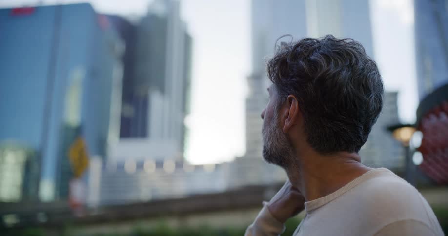 A grayhaired bearded man gazes upward with a thoughtful expression amid towering glass skyscrapers the soft daylight and blurred city backdrop emphasizing a quiet moment of reflection