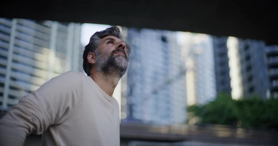 A middleaged man with saltandpepper hair and a beard looks upward toward the sky he is standing outdoors in an urban environment framed by tall city buildings in the background