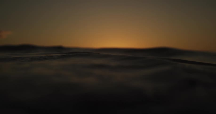 A softly blurred ocean surface undulates in the foreground as a warm amber glow from the setting sun lingers on the horizon creating a calm cinematic seascape where low waves catch the fading light