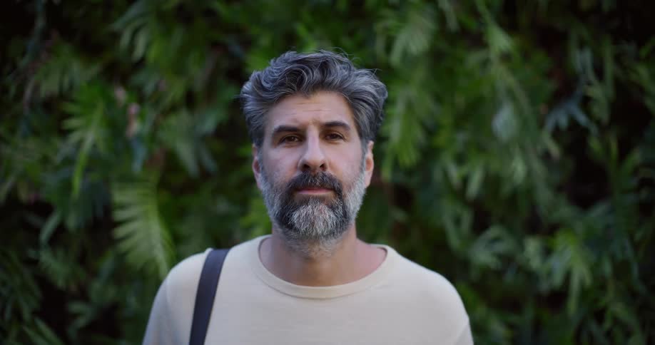 A middleaged man with gray and dark facial hair stands in front of a tree at midday adjusting a strap on his shoulder with a sad expression