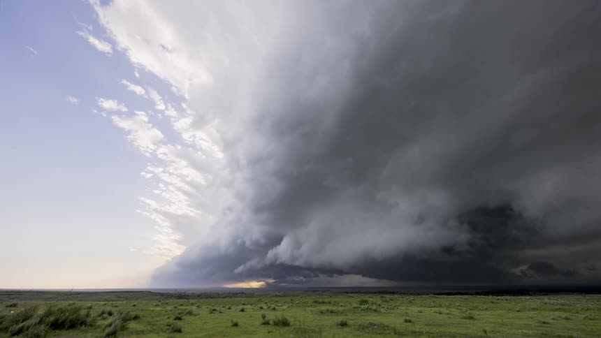 Massive storm clouds roll over a flat green grassland under a partly cloudy sky