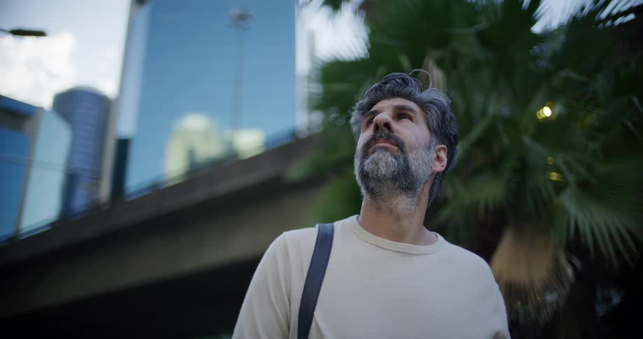 A man with graying hair and a beard stands outdoors in a city looking upward toward the tall buildingsthe scene is captured in a lowangle shot with palm trees and a modern glass skyscraper visible in the background