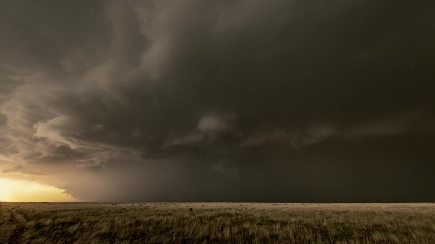 A vast grassland stretches beneath an immense dark storm cloud with a faint glow of sunlight on the horizon heightening the dramatic contrast between calm land and approaching severe weather