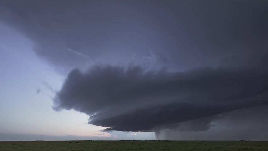A massive layered storm cloud looms low over an open plain at dusk its dark rotating form trailing curtains of rain and lightning as it dominates the horizon with an ominous cinematic presence