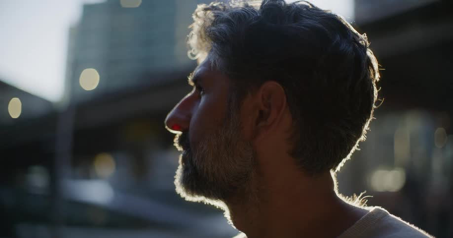 A middleaged man with a graying beard stands outdoors during sunset looking toward the sky and then turning his head to the side