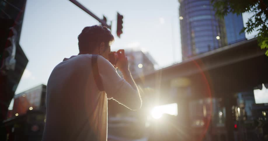 Man photographs an urban scene in backlight near an intersection with modern buildings and traffic lights in the background