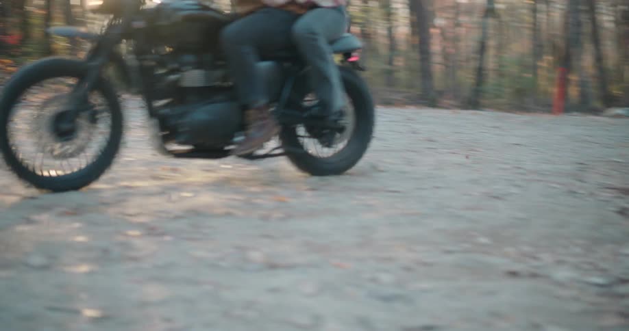 A motorcycle carrying two riders travels down a paved road through a forest with autumn leavesthe bike speeds toward the camera and then leans into a sharp turn before disappearing out of the frame